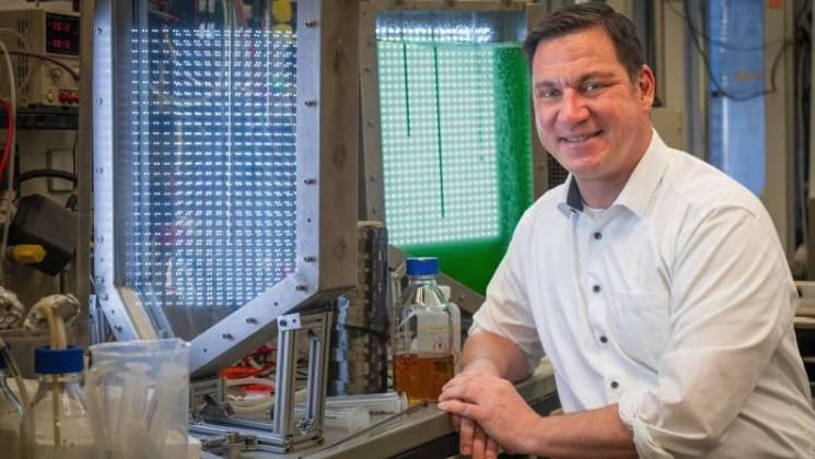 Man in a lab coat smiling next to scientific equipment.