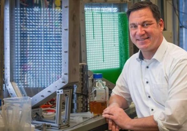 Man in a lab coat smiling next to scientific equipment.